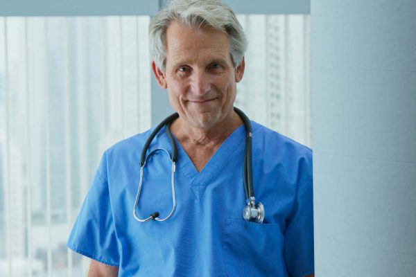 Portrait of Senior Caucasian male nurse or doctor looking at camera while standing in hospital. Trustworthy medical professional in blue scrubs with stethoscope smiling in clinic