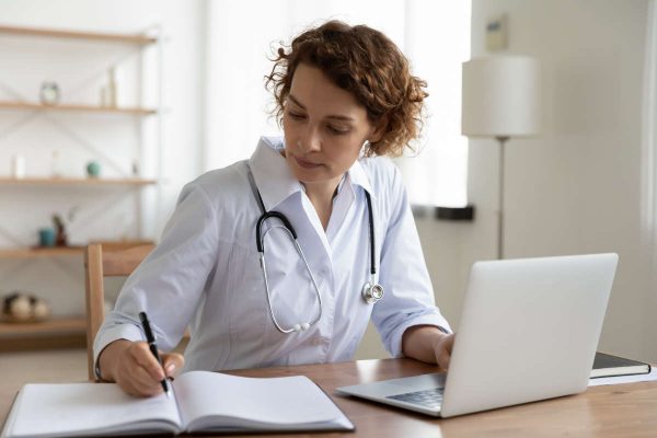Serious,Female,Doctor,Using,Laptop,And,Writing,Notes,In,Medical