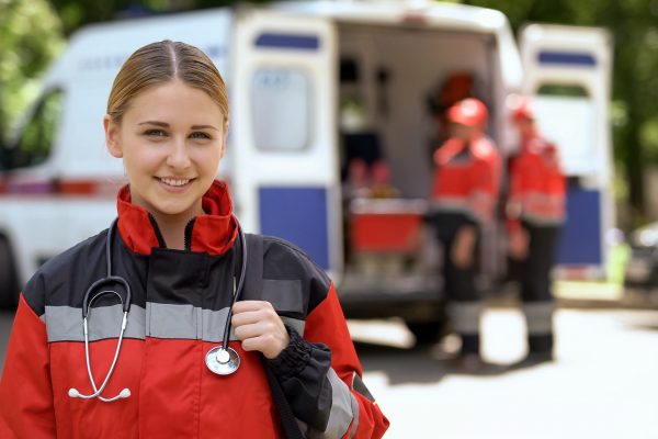 Female,Paramedic,Smiling,Into,Camera,,Ambulance,Crew,Blurred,On,Background